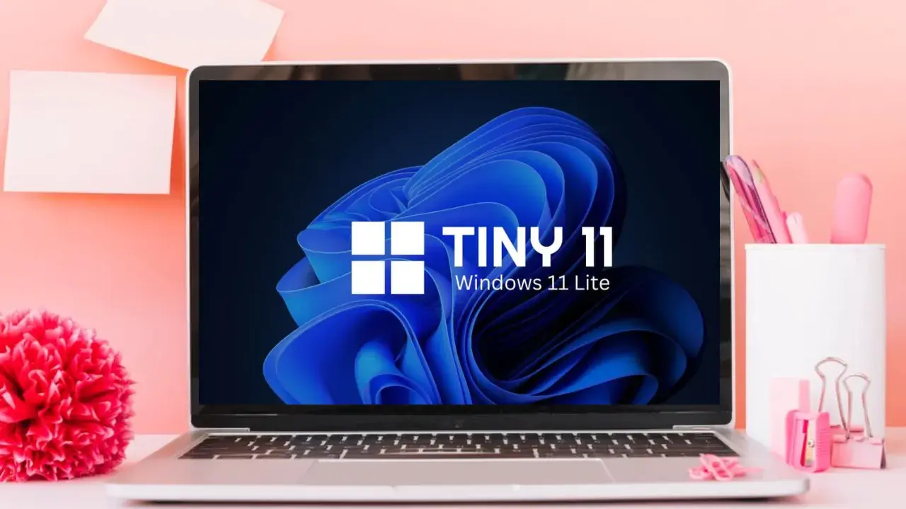 Laptop mit "TINY 11 Windows 11 Lite" auf dem Bildschirm, umgeben von rosa Büroartikeln und einer roten Blume.
