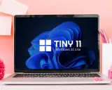 Laptop mit "TINY 11 Windows 11 Lite" auf dem Bildschirm, umgeben von rosa Büroartikeln und einer roten Blume.