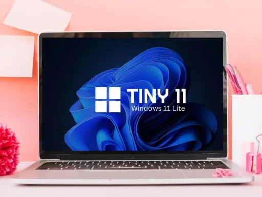 Laptop mit "TINY 11 Windows 11 Lite" auf dem Bildschirm, umgeben von rosa Büroartikeln und einer roten Blume.